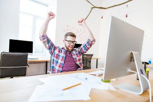 Happy successful man in glasses with raised hands sitting on workplace in office