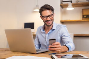 Young happy businessman smiling while reading his smartphone. Portrait of smiling business man reading message with smartphone in office. Man working at his desk at office.