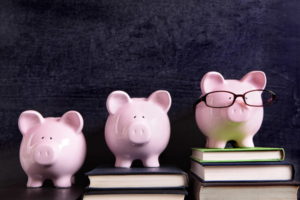 Three pink piggy banks standing on books next to a blackboard with retirement savings message. Sharp focus on the piggy banks.