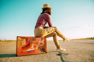 Traveler woman sits on retro suitcase and looks away on road. Suitcase with stamps flags representing each country traveled.