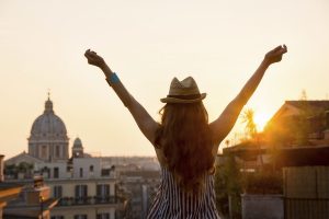 Seen from behind, a woman is standing with outstretched arms, looking out at the city of Rome at sunset in summer.