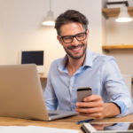 Young happy businessman smiling while reading his smartphone. Portrait of smiling business man reading message with smartphone in office. Man working at his desk at office.