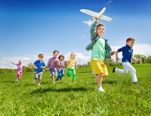 Group of active running kids with boy holding big white airplane toy in the field during summer sunny day