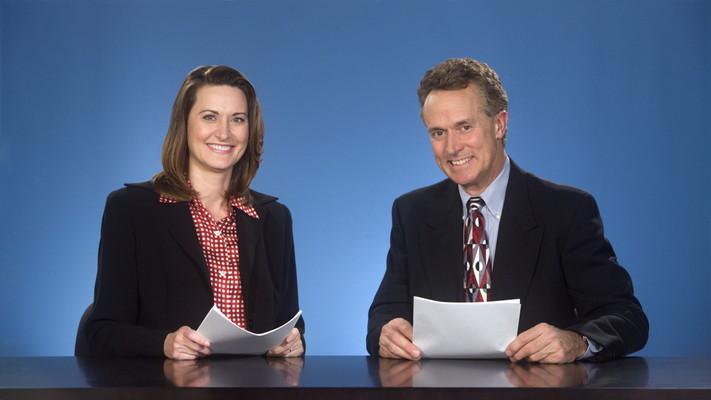 Male and female newcasters sitting at desk smiling at viewer.