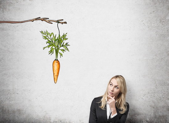 young pretty woman with her hand on chin looking up at a painted carrot tied to a branch. Concrete background. Front view. Concept of reward.